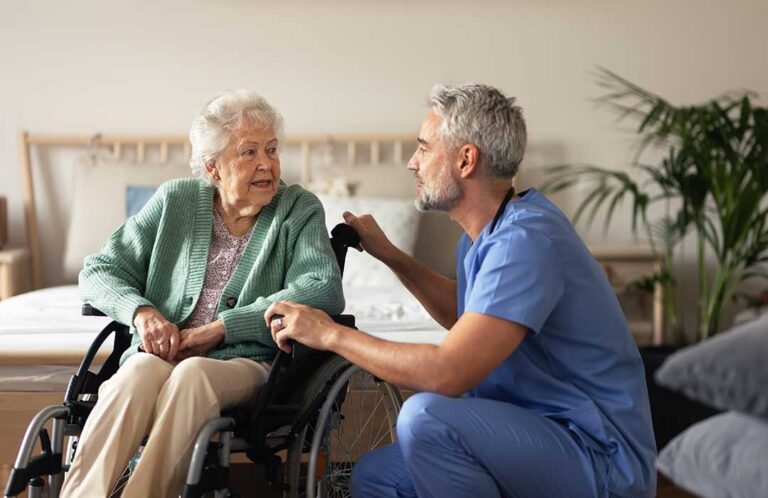 Caregiver assisting an older adult in a bright assisted living apartment with a walker nearby.