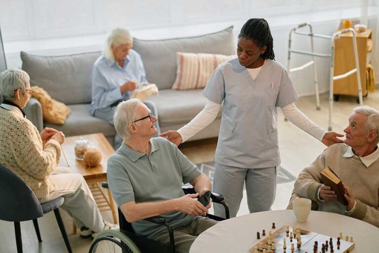 An older adult sits at a kitchen table sorting medication and paperwork, with sunlight highlighting everyday details.